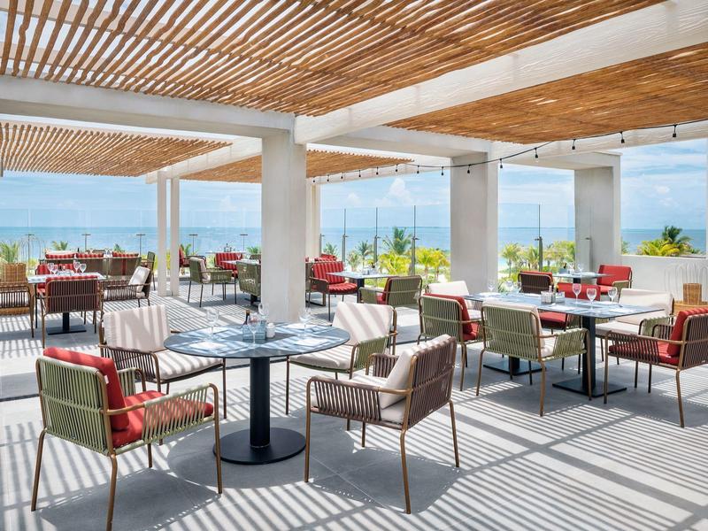 Área de restaurante abierta con mesas y sillas, mesas puestas y vista al mar bajo techo de listones de madera.