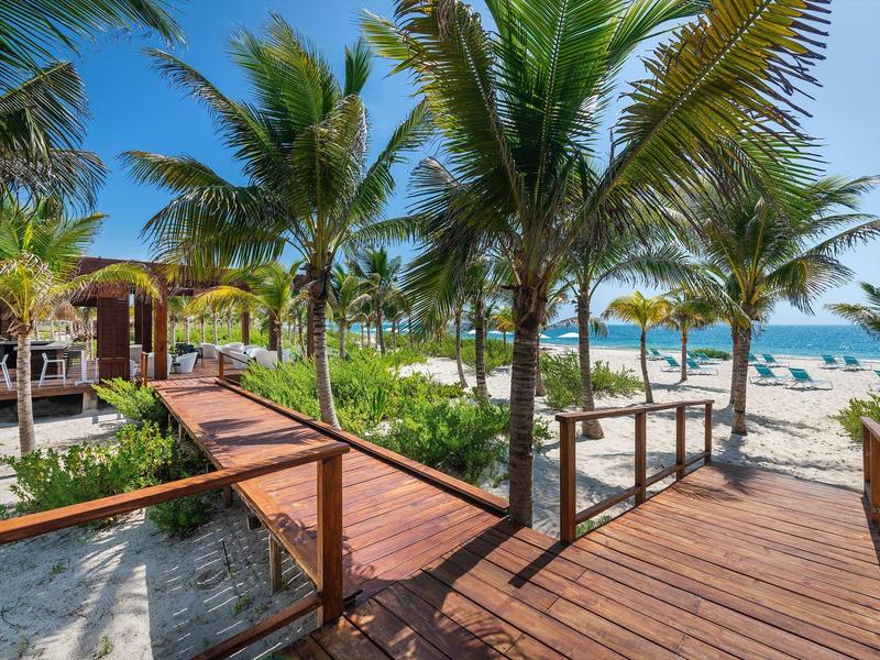 Pasarela de madera entre palmeras que conduce a la playa con tumbonas y cielo despejado.