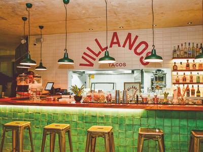 Bar moderna con sgabelli verdi e illuminazione calda, decorata con insegna Villano Taco.