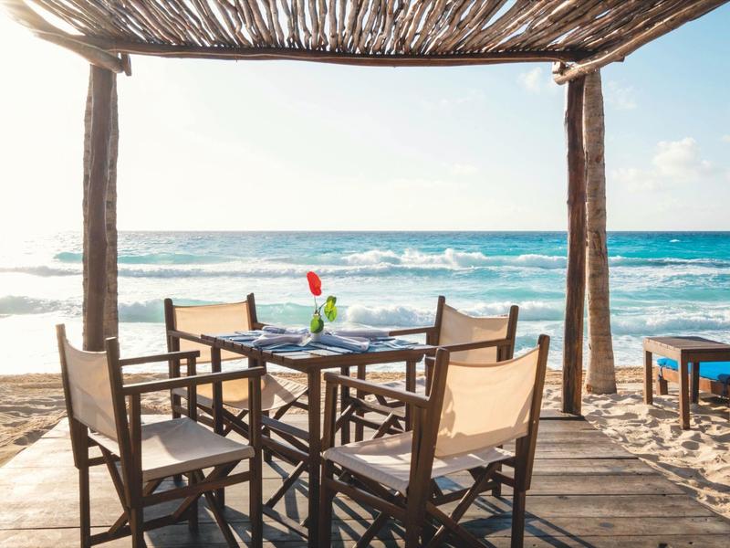 Terrazza con tavolo e sedie in legno sulla spiaggia con vista sul mare sotto un tetto di paglia.