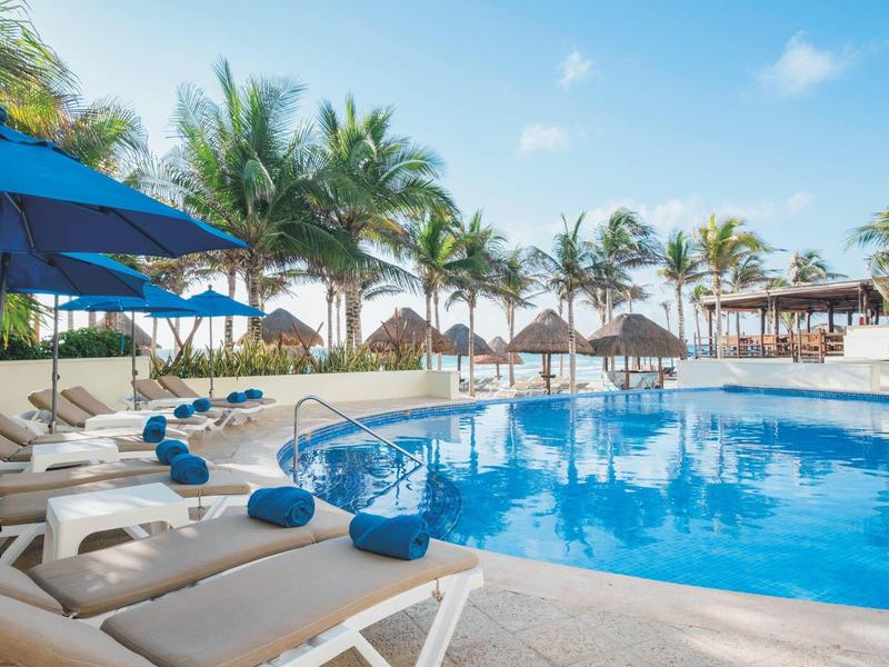 Zona piscina con lettini, asciugamani blu e ombrelloni, circondata da palme.