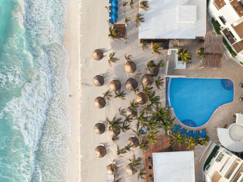 Vista aerea di un hotel con piscina, palme e spiaggia con onde.