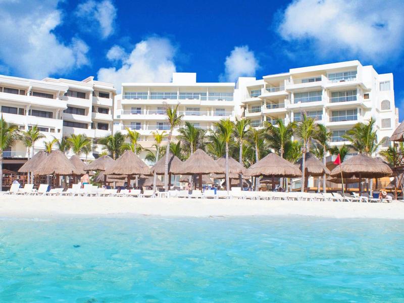 Hotel moderno sulla spiaggia con facciata bianca, palme e mare azzurro cristallino in primo piano.