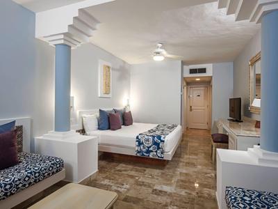 Habitación de hotel con decoración azul, cama doble, sofá y piso de madera clara.