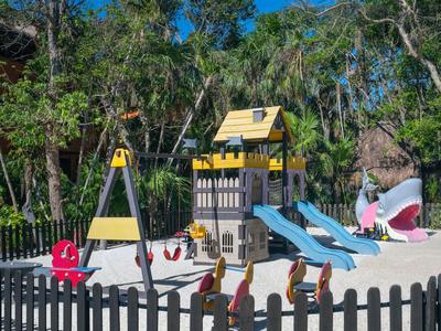 Área de juegos infantil colorida con tobogán y columpios, rodeada de árboles y cerca al aire libre.