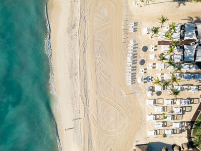 Vista aérea de una playa con agua clara, arena y sombrillas junto a la piscina del hotel.
