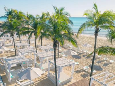 Ein sonniger Strand mit Palmen und weißen Liegen neben dem blauen Meer.