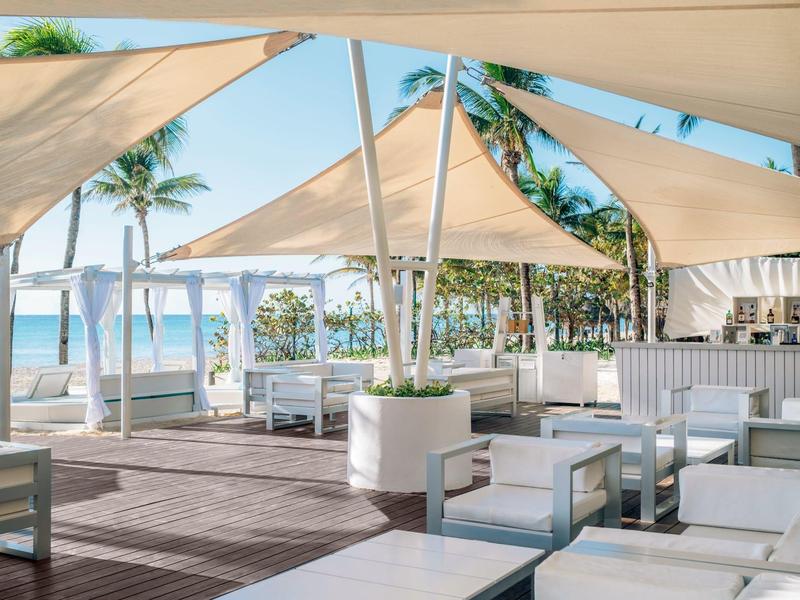 Lounge moderna al aire libre con muebles blancos y vista al mar bajo toldos de vela.