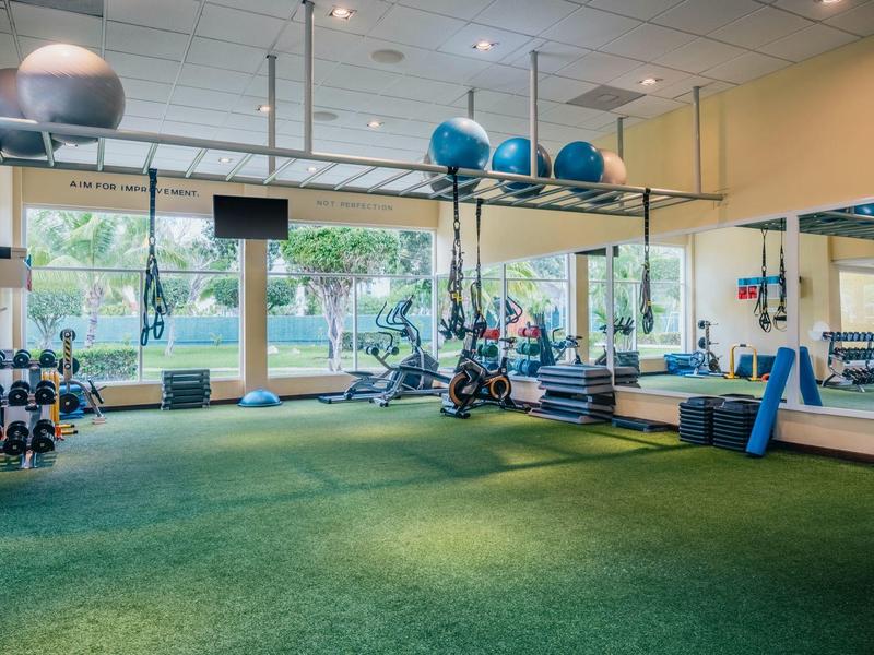 Gimnasio luminoso con gran espejo, suelo verde y varios equipos de ejercicio.