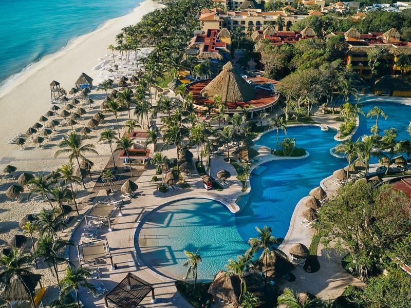 Vista aérea de un resort con playa, piscina, tumbonas y palmeras junto al océano.