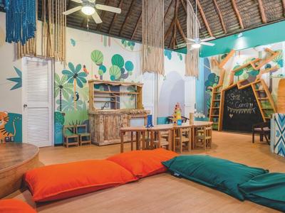 Buntes Kindergartenzimmer mit Holzmöbeln, Wandmalereien und großen Sitzkissen auf dem Boden.