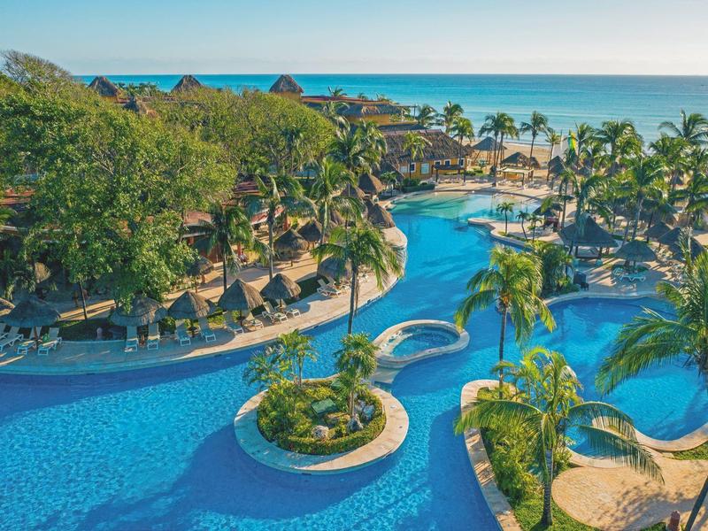 Tropical hotel pool with palm trees and lake in the background under blue sky.