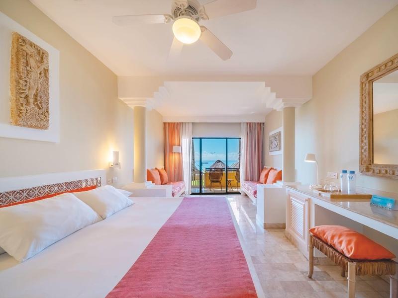Bright hotel room with large bed, seating area, and sea view through large windows.