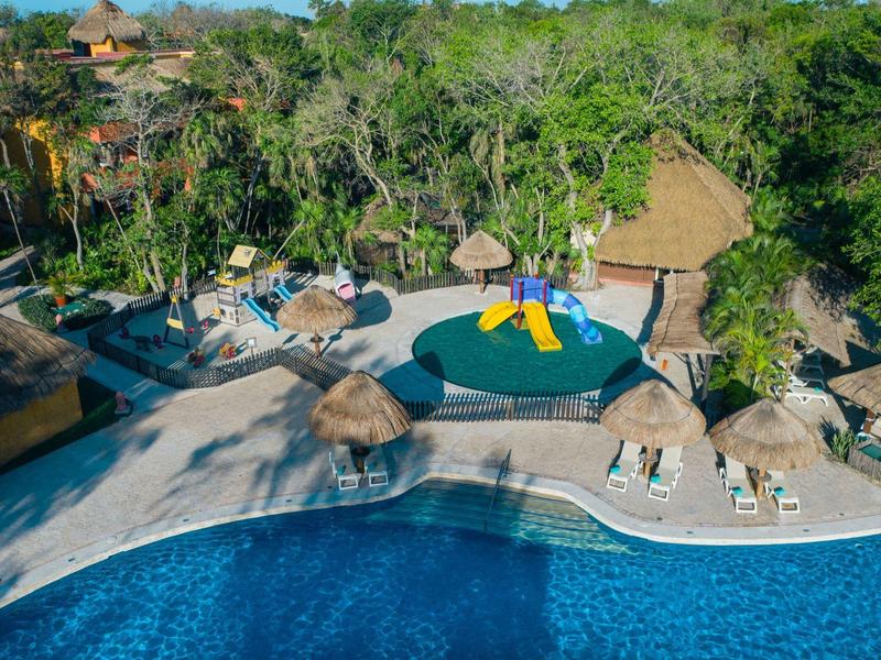 View of a pool area with a kids' pool, water slide, and sun loungers surrounded by trees.