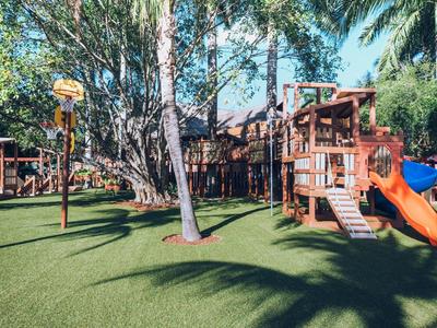 Kinderspielplatz mit Holzgerüsten, Rutsche, Kletterwand und künstlichem Gras unter Palmen.