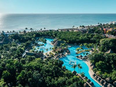 Luftaufnahme eines Resorts mit türkisblauem Pool, viel Grün und Blick auf das Meer am Horizont.