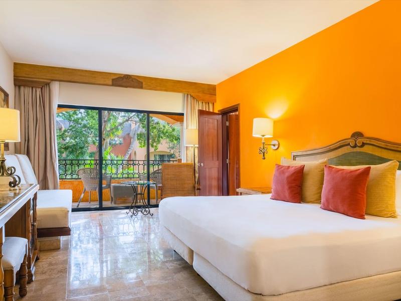 Camera d'hotel luminosa con un grande letto, cuscini colorati e accesso a un balcone.