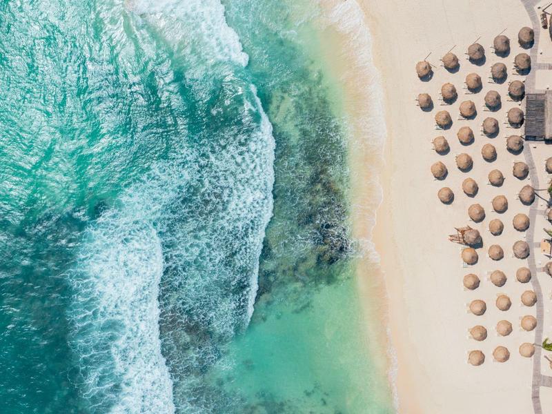 Vista aerea di una spiaggia con acqua turchese, onde e file di ombrelloni.
