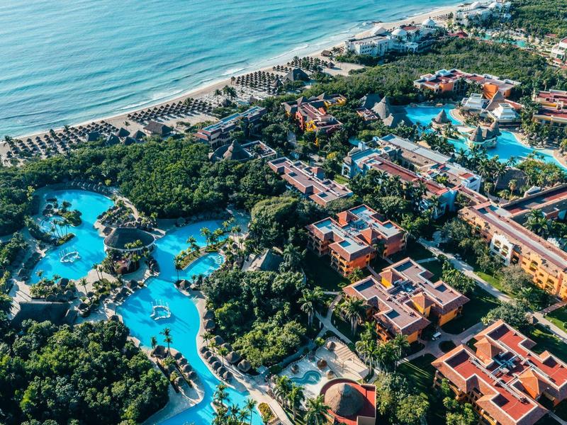 Vista aerea di un resort costiero con più piscine, vegetazione rigogliosa e una spiaggia sul mare.
