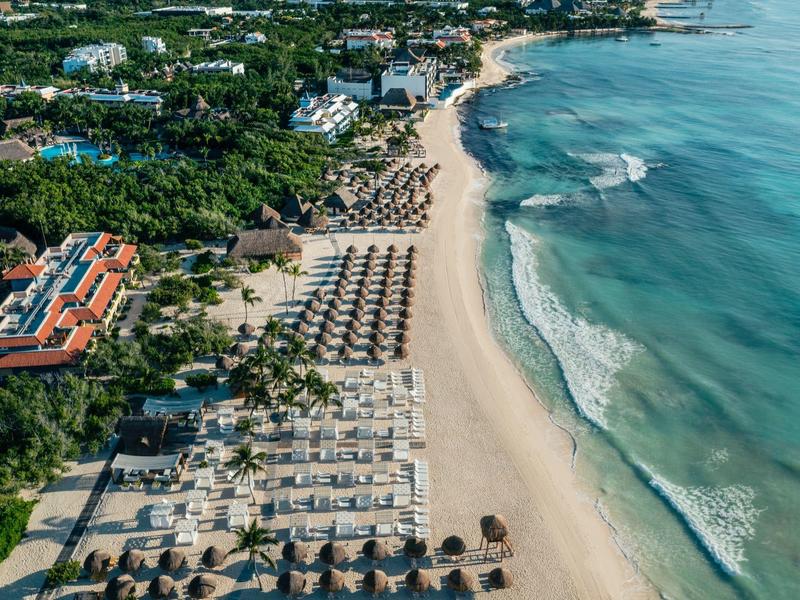 Vista aerea di una larga spiaggia sabbiosa con file di ombrelloni e lettini accanto a un hotel.