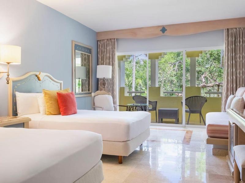 Camera d'hotel luminosa con due letti, zona salotto e vista su una terrazza con giardino.