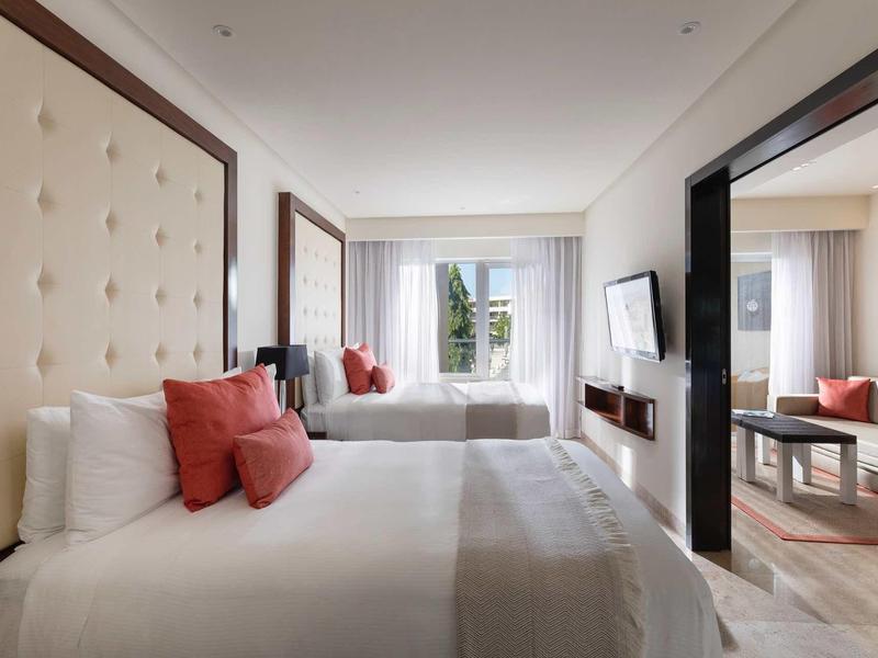 Camera d'hotel luminosa con due letti, testiera imbottita e TV a parete.