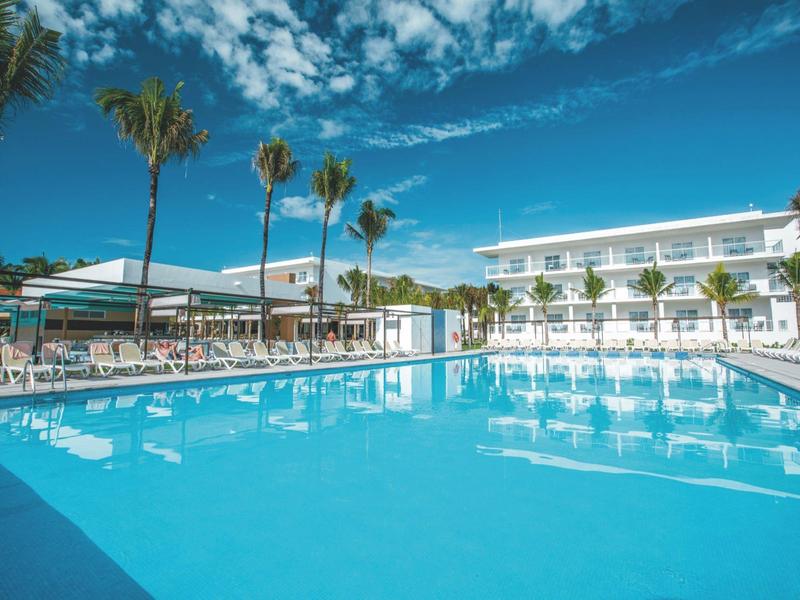 Large pool with clear blue water in front of a white hotel under a bright blue sky.