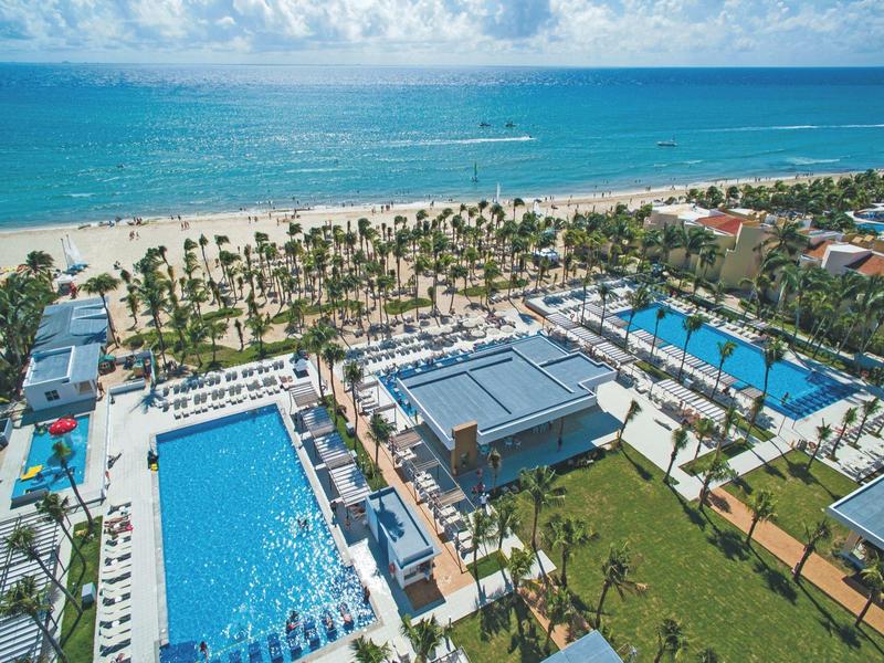 Large resort with multiple pools, palm trees, and beach by the sea on a sunny day.