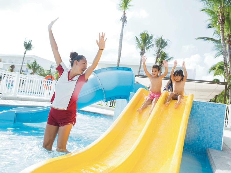 Children slide down yellow water slide while supervisor watches in swimming pool.