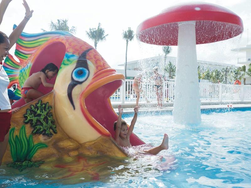 Children sliding down a colorful fish slide into a pool with a red mushroom sprinkler.
