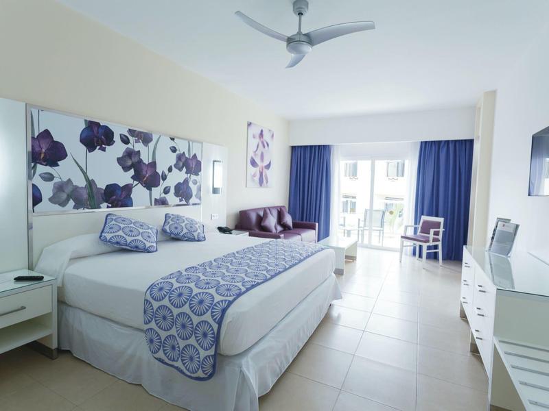 Bright hotel room with large bed, blue curtains, and modern decor.