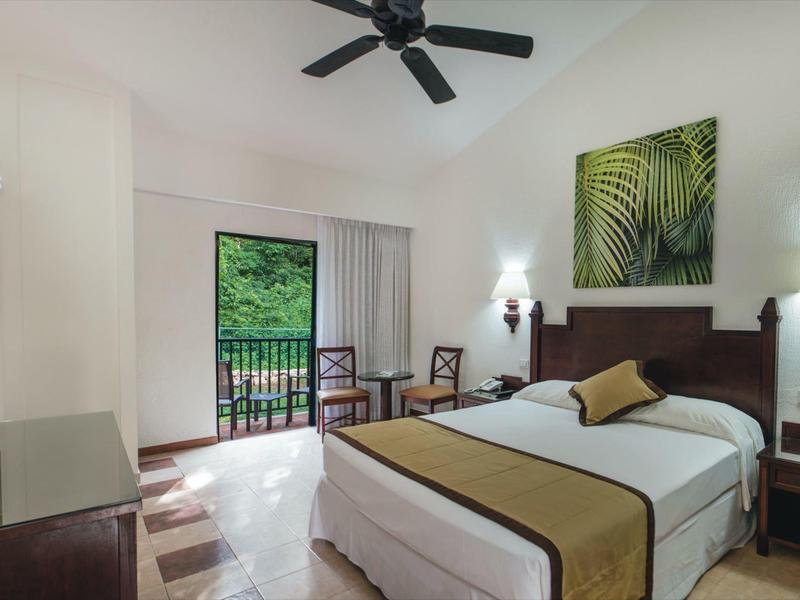 Camera d'hotel luminosa con letto matrimoniale, balcone e arredi moderni con ventilatore a soffitto.