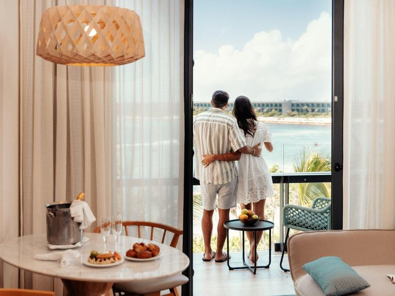 Paar steht auf Balkon, schaut aufs Meer, gemütlicher Raum mit hellem Holz und sanften Farben.