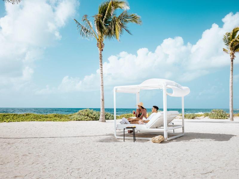 Zwei Personen entspannen auf einem weißen Strandbett unter blauem Himmel mit Palmen am Meer.