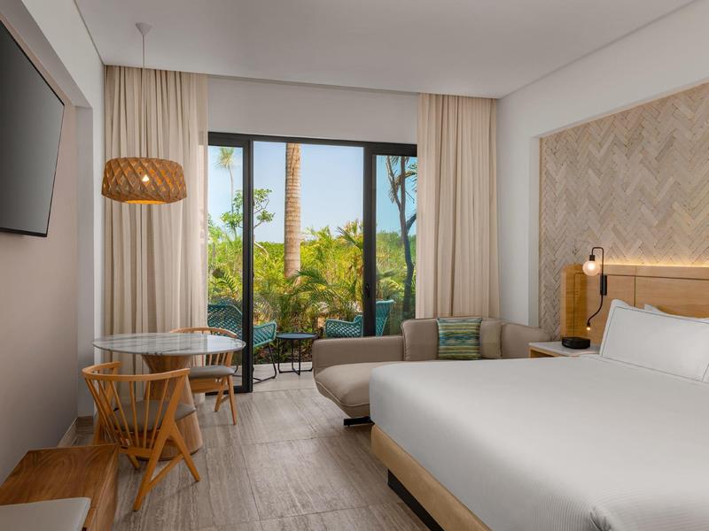 Helles Zimmer mit Doppelbett, Sitzbereich, Holzmöbeln und großem Fenster mit Blick auf Garten.