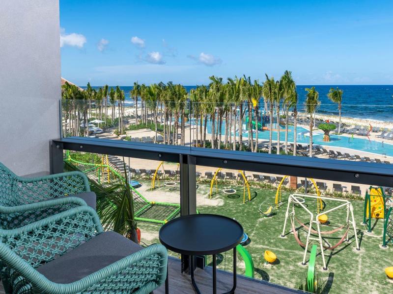 Balkon mit grünen Stühlen und Tisch, Blick auf Palmen, Spielplatz und Meer unter blauem Himmel.