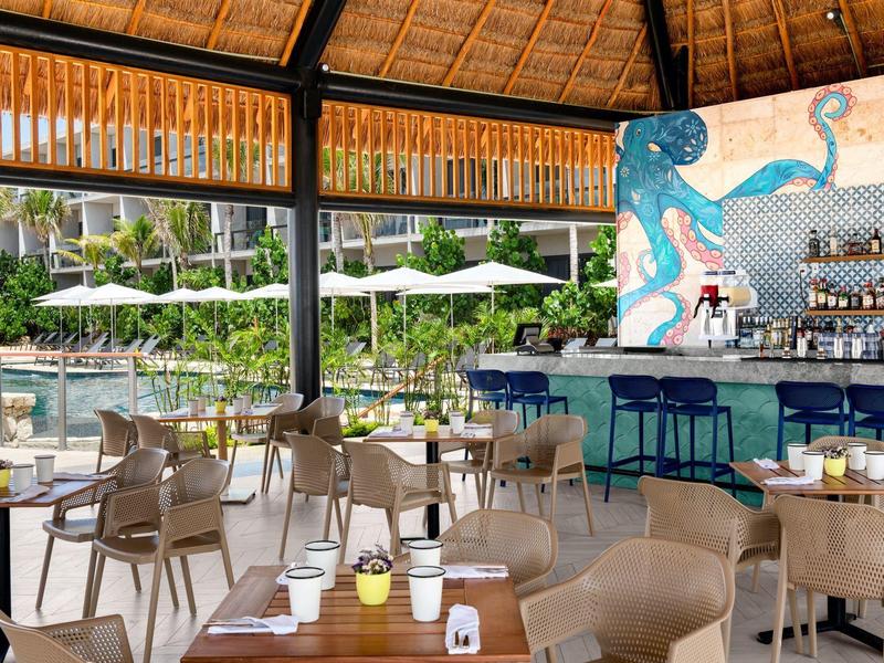Offenes Restaurant mit Holzstuhlgruppen, blauem Barbereich und Blick auf Pool mit grüner Umgebung.