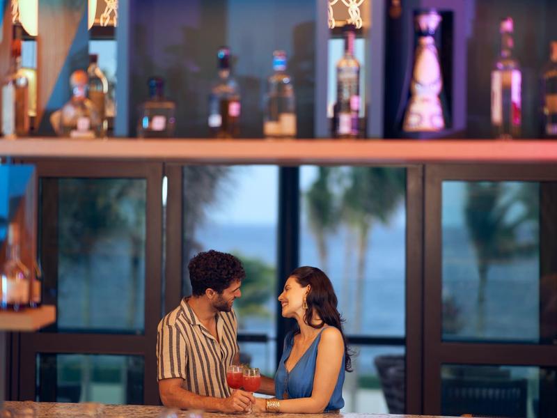 Ein Paar sitzt lächelnd am Bar-Tresen, vor großen Fenstern mit Palmenblick bei Abendlicht.
