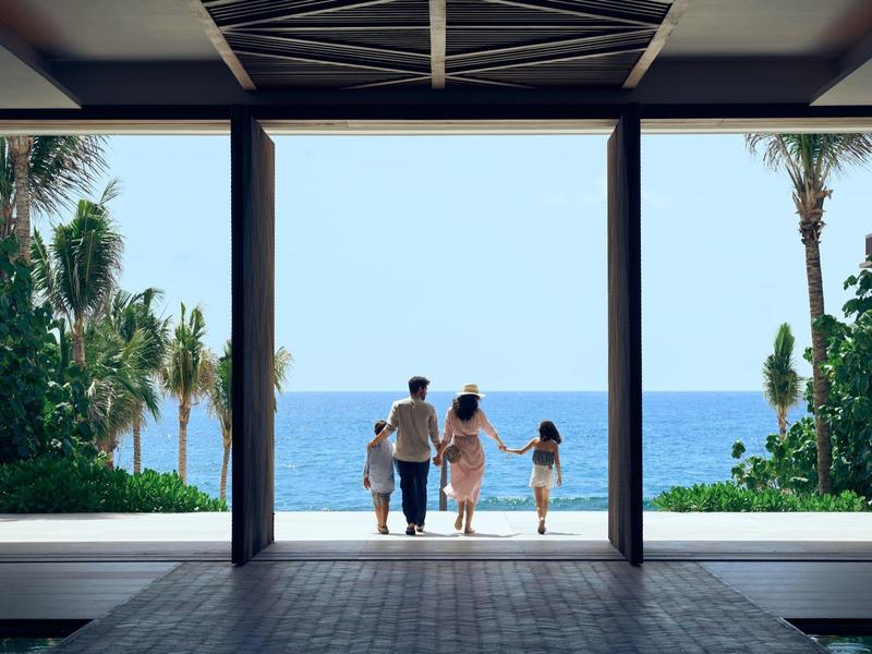 Familie mit zwei Kindern steht am offenen Durchgang, Blick auf Meer und Palmen bei klarem Himmel.
