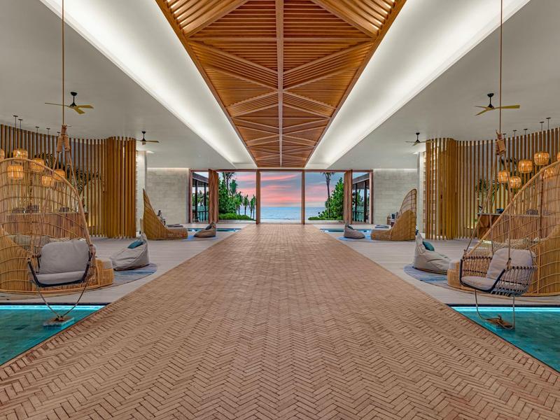 Großer, offener Lobbybereich mit Holzdach, Sitzgruppen, Pflanzen und Blick auf Meer und Sonnenuntergang.