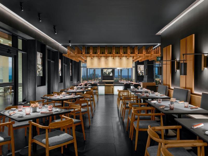 Modernes Restaurant mit Holzstühlen, grauen Wänden, langen Tischen und stilvoller Beleuchtung.