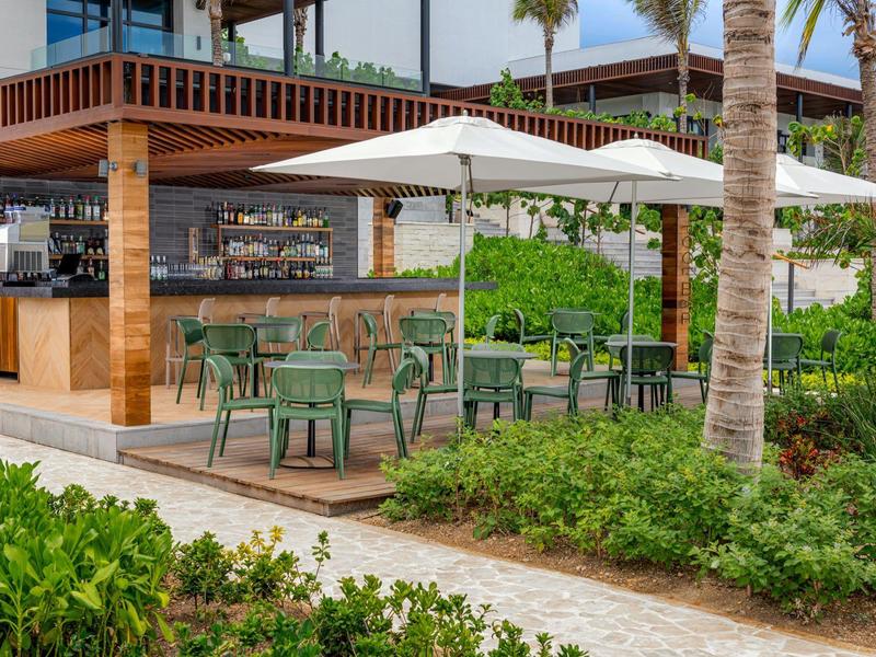 Modernes Café mit Holzstruktur, grünen Stühlen, weißen Sonnenschirmen und Palmen im Außenbereich.