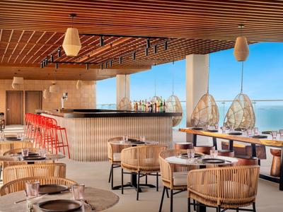 Restaurant d'hôtel moderne avec plafond en bois, chaises en osier et vue sur la mer.