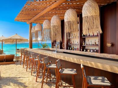 Bar de plage avec comptoir en bois, tabourets en osier et léger auvent au bord de la mer sous un ciel dégagé.