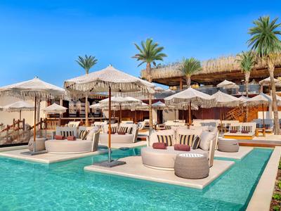 Zone de piscine de luxe avec parasols, transats et palmiers sous un ciel clair.