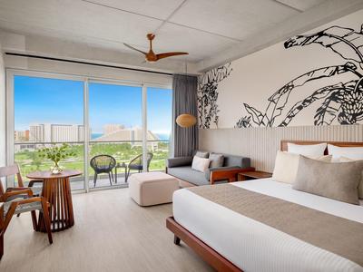 Chambre d'hôtel lumineuse avec grand lit, œuvre murale, coin salon et balcon avec vue sur la ville.