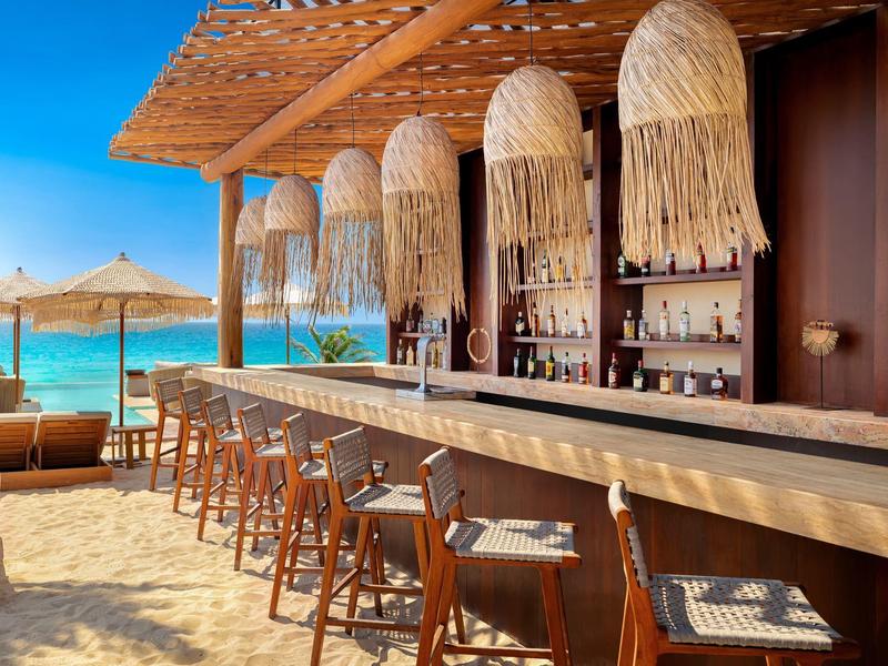 Bar de plage avec comptoir en bois, tabourets en osier et léger auvent au bord de la mer sous un ciel dégagé.