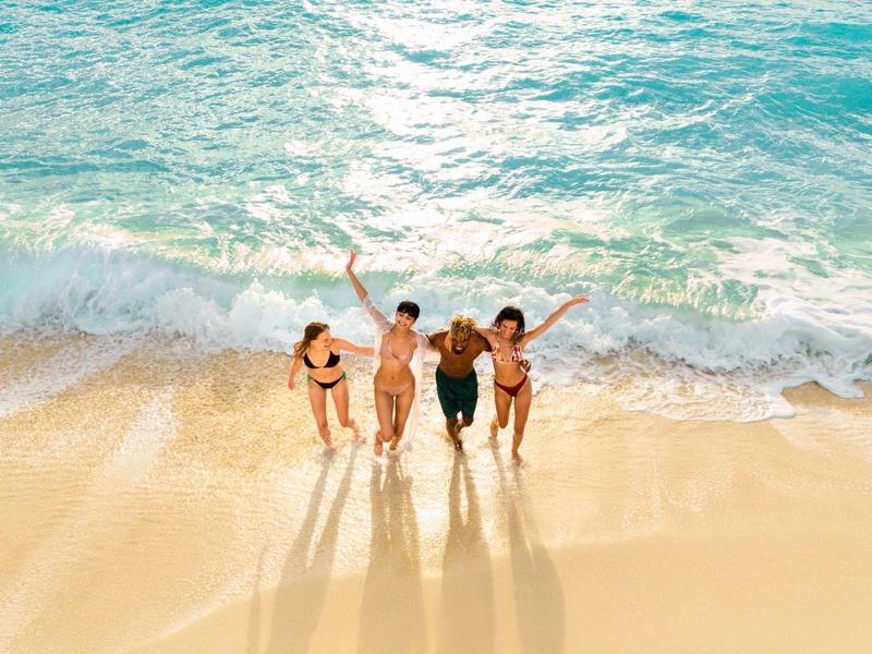 Quatre personnes marchant le long de la plage ensoleillée, mer et vagues en arrière-plan.