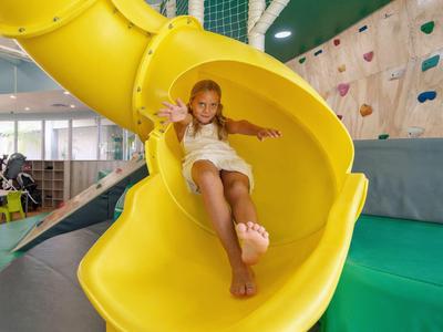 Ein Mädchen rutscht lachend eine große gelbe Rutschbahn in einer bunten Indoor-Spielhalle hinunter.