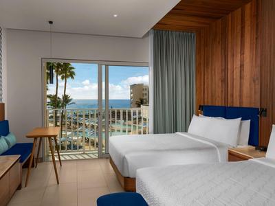 Hotelzimmer mit zwei Betten, Holzwänden, Balkon und Blick auf Meer und Palmen.